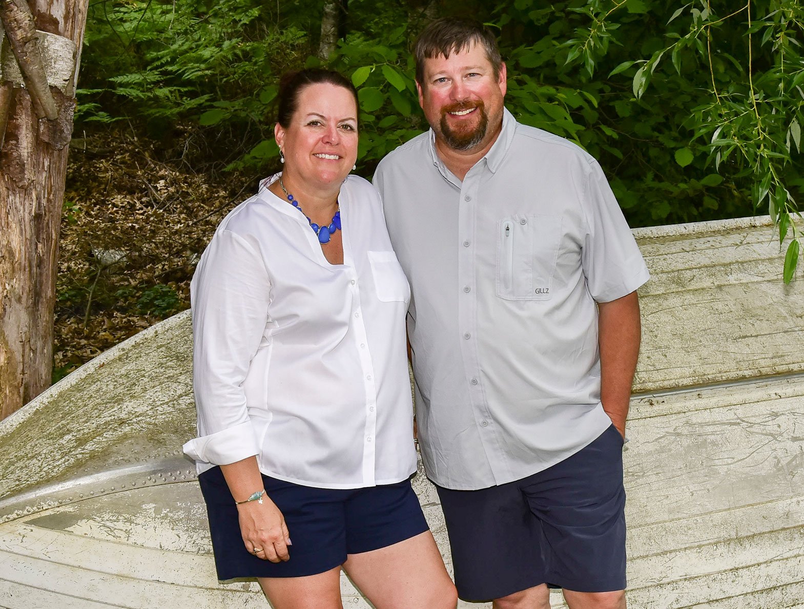 A woman and a man standing outdoors in front of an old boat and green foliage, smiling at the camera.