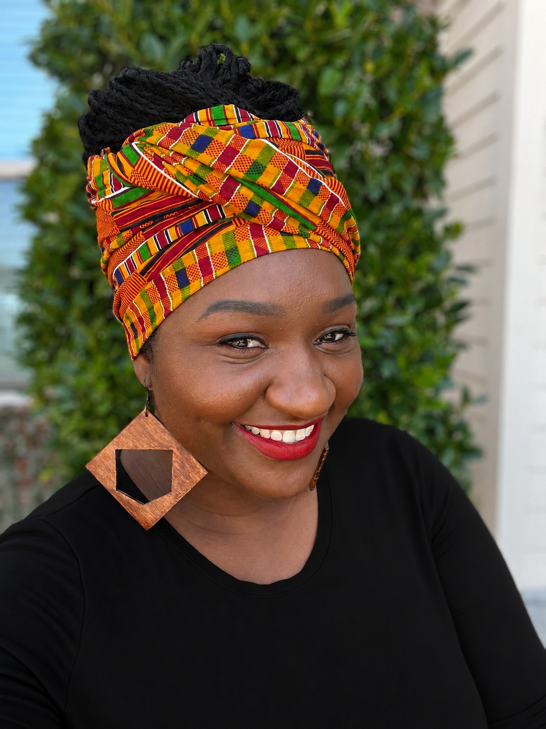 A woman smiling outdoors, wearing a colorful patterned headwrap, large geometric wooden earrings, and a black top. Green foliage and a portion of a house are visible in the background.
