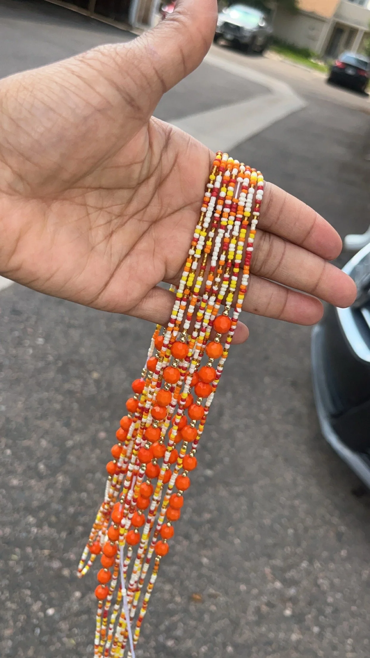 A hand holding multiple strands of colorful beaded jewelry with orange, yellow, white, and red beads, against a background of a street with parked cars and residential buildings.