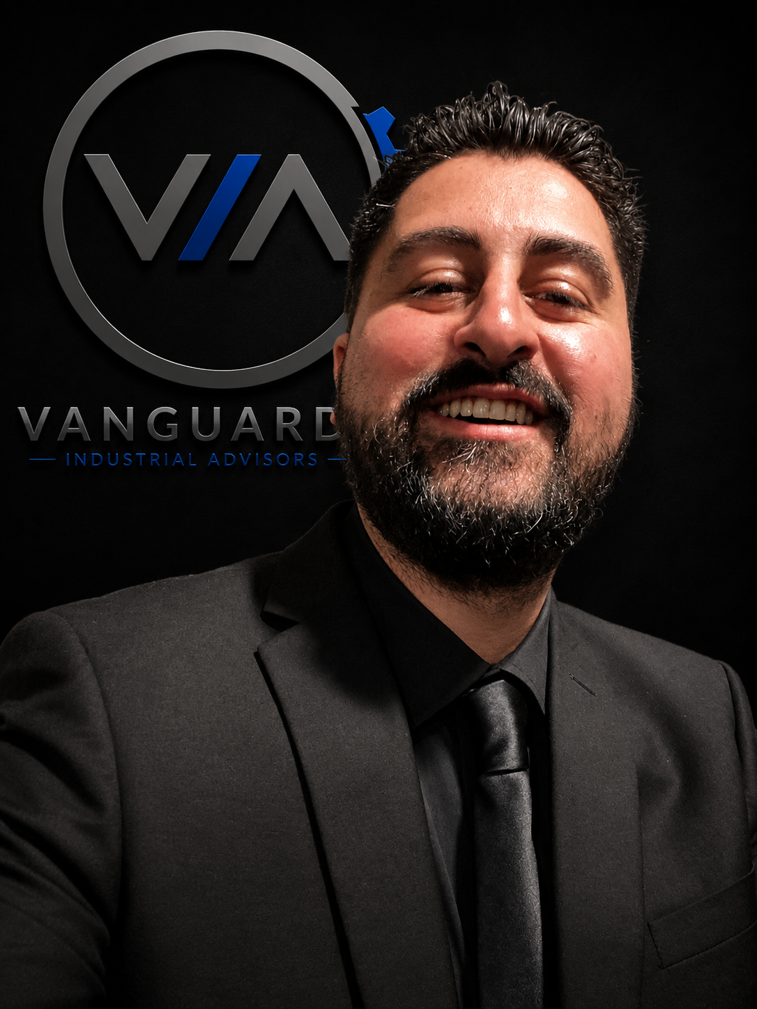 Man in a black suit and tie taking a selfie in front of a Vanguard logo with the text "VANGUARD INDUSTRIAL ADVISORS" on a black background.