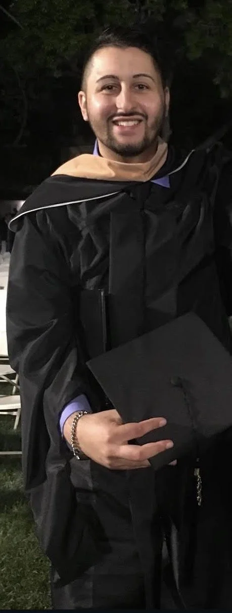 A young man in a black graduation gown holding a black mortarboard cap is smiling at the camera. He is outdoors at night, with trees or bushes in the background.