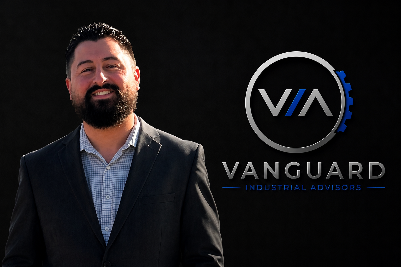 A smiling man with dark hair, a beard, wearing a business suit and shirt, standing next to a sign that reads "Vanguard Industrial Advisors" with a logo.