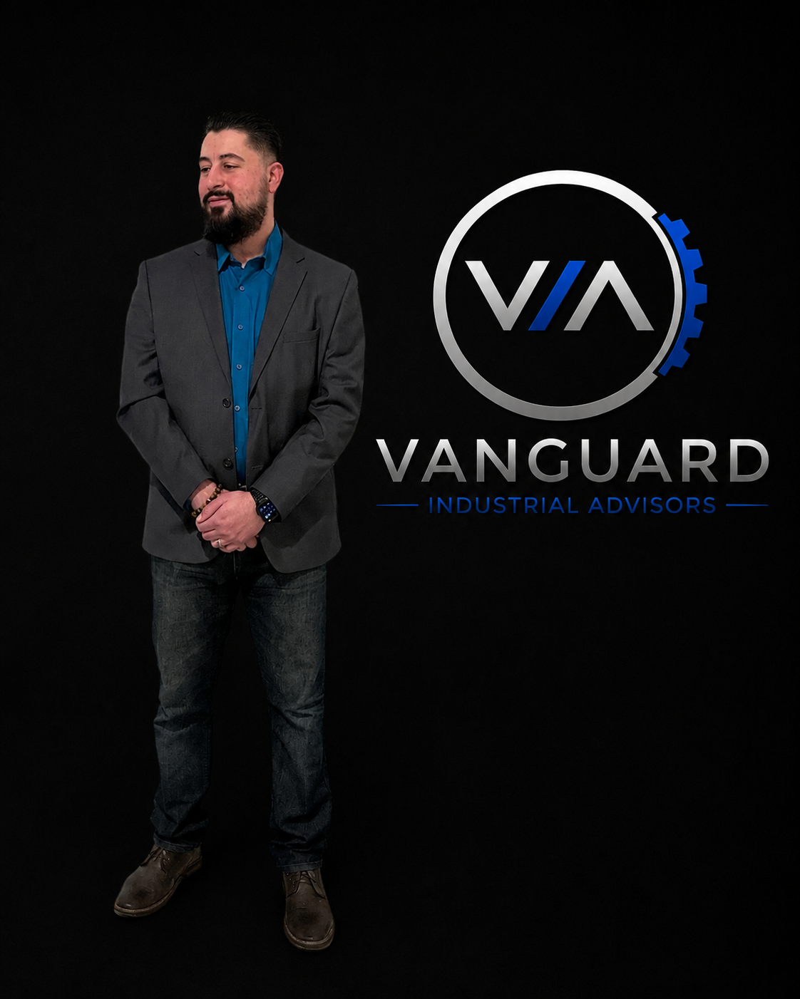 A man with a beard wearing a gray blazer, blue shirt, dark jeans, and brown shoes standing against a black background with a 'Vanguard Industrial Advisors' logo.