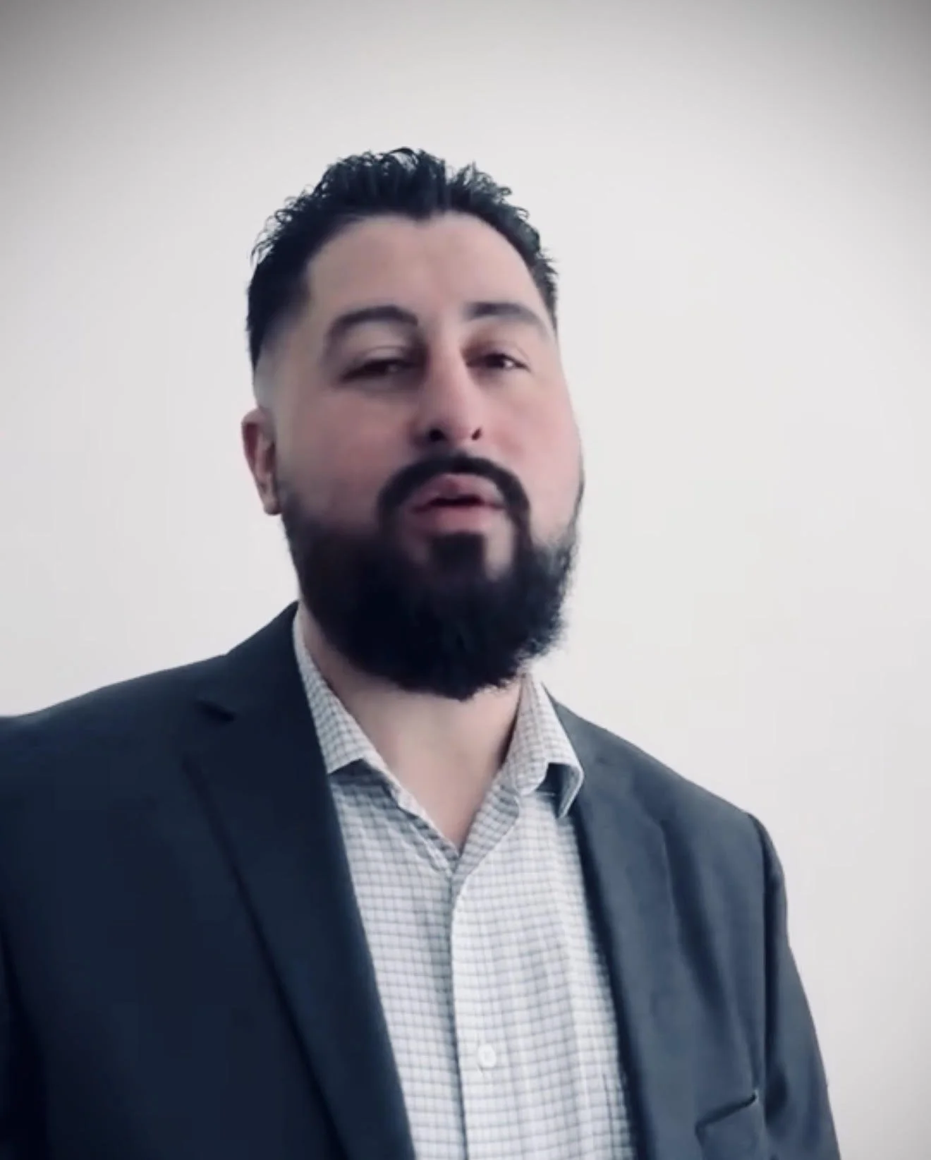 A man with dark hair, a beard, and a mustache wearing a suit jacket and checkered shirt stands against a plain white background.