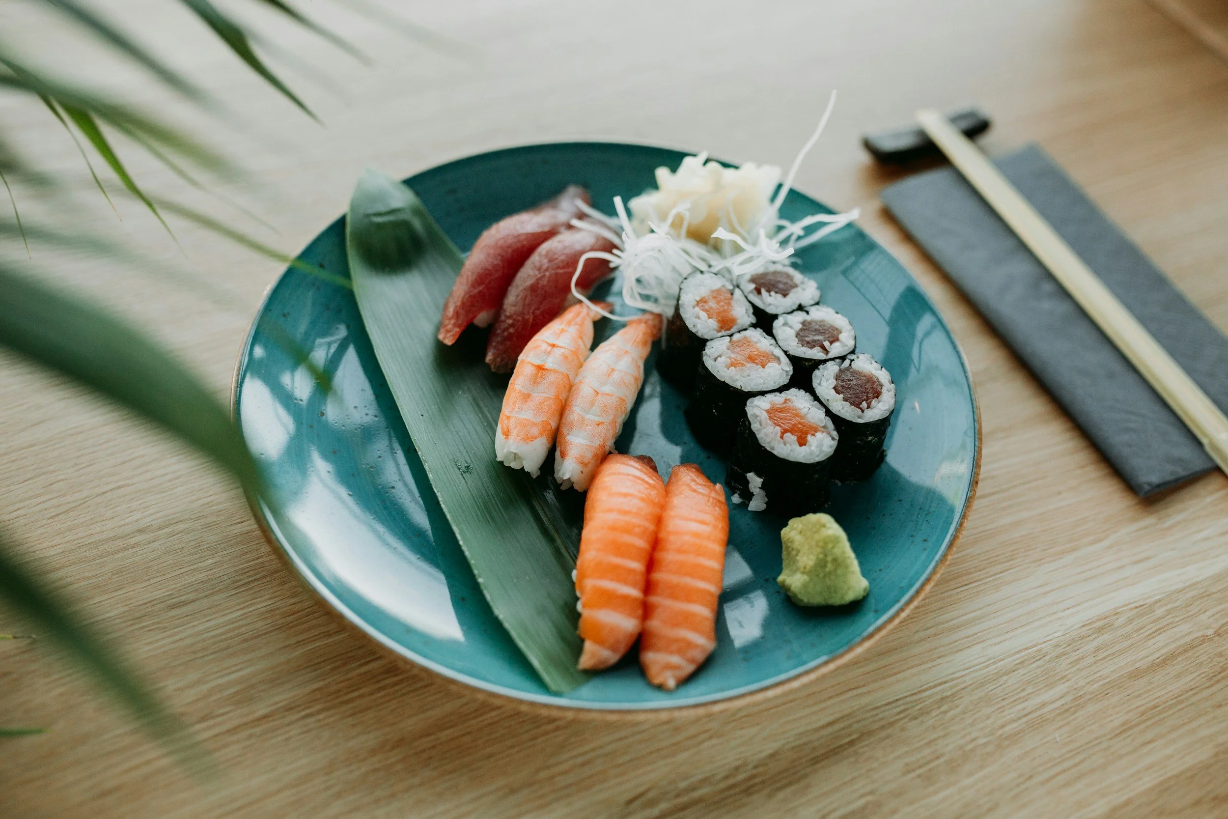 Sushi platter with various types of sushi, including salmon, shrimp, and tuna rolls, on a green leaf on a blue plate, with wasabi and shredded daikon, on a wooden table.