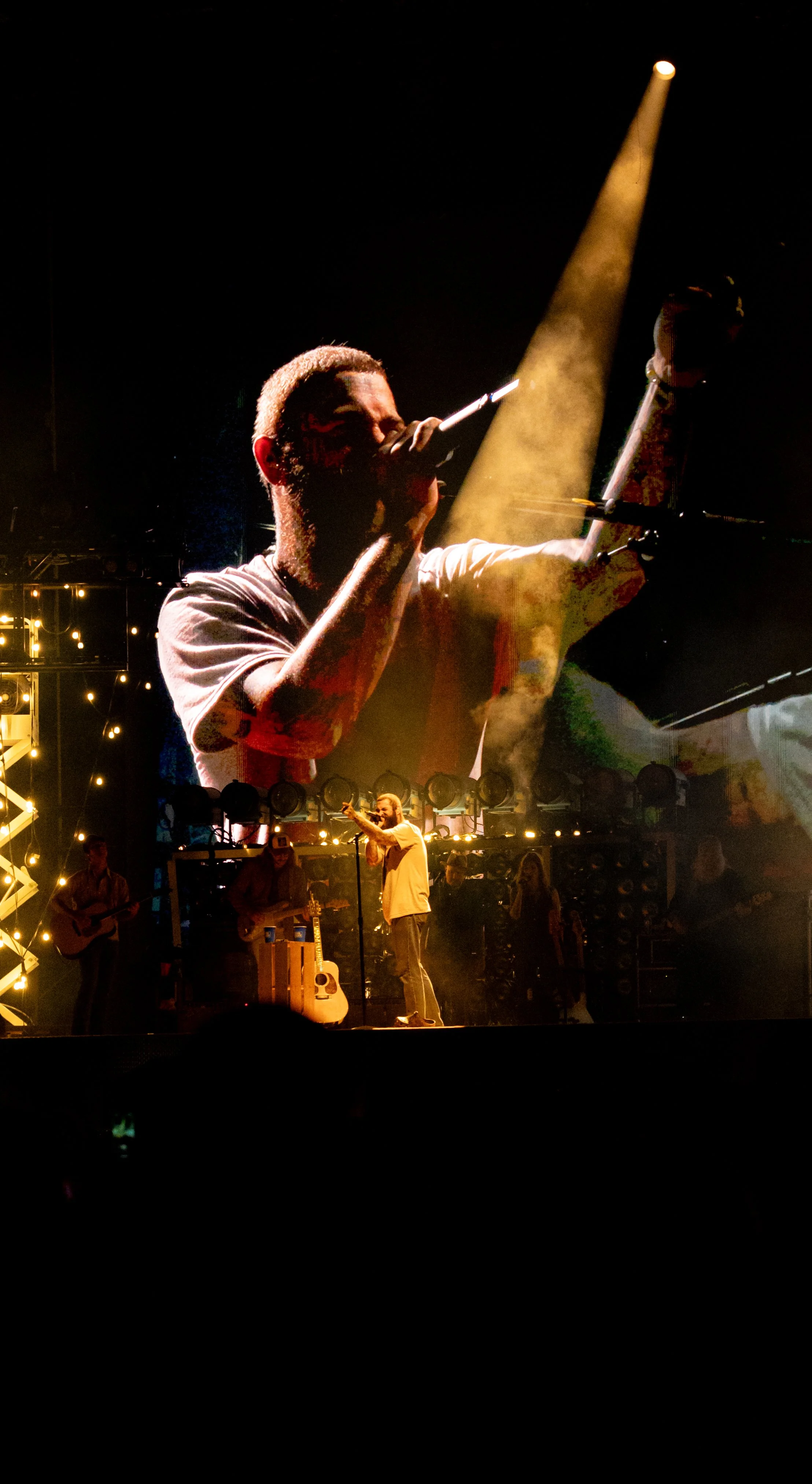 A live music concert with a performer singing into a microphone on stage, illuminated by yellow and white stage lights, with band members playing instruments in the background.