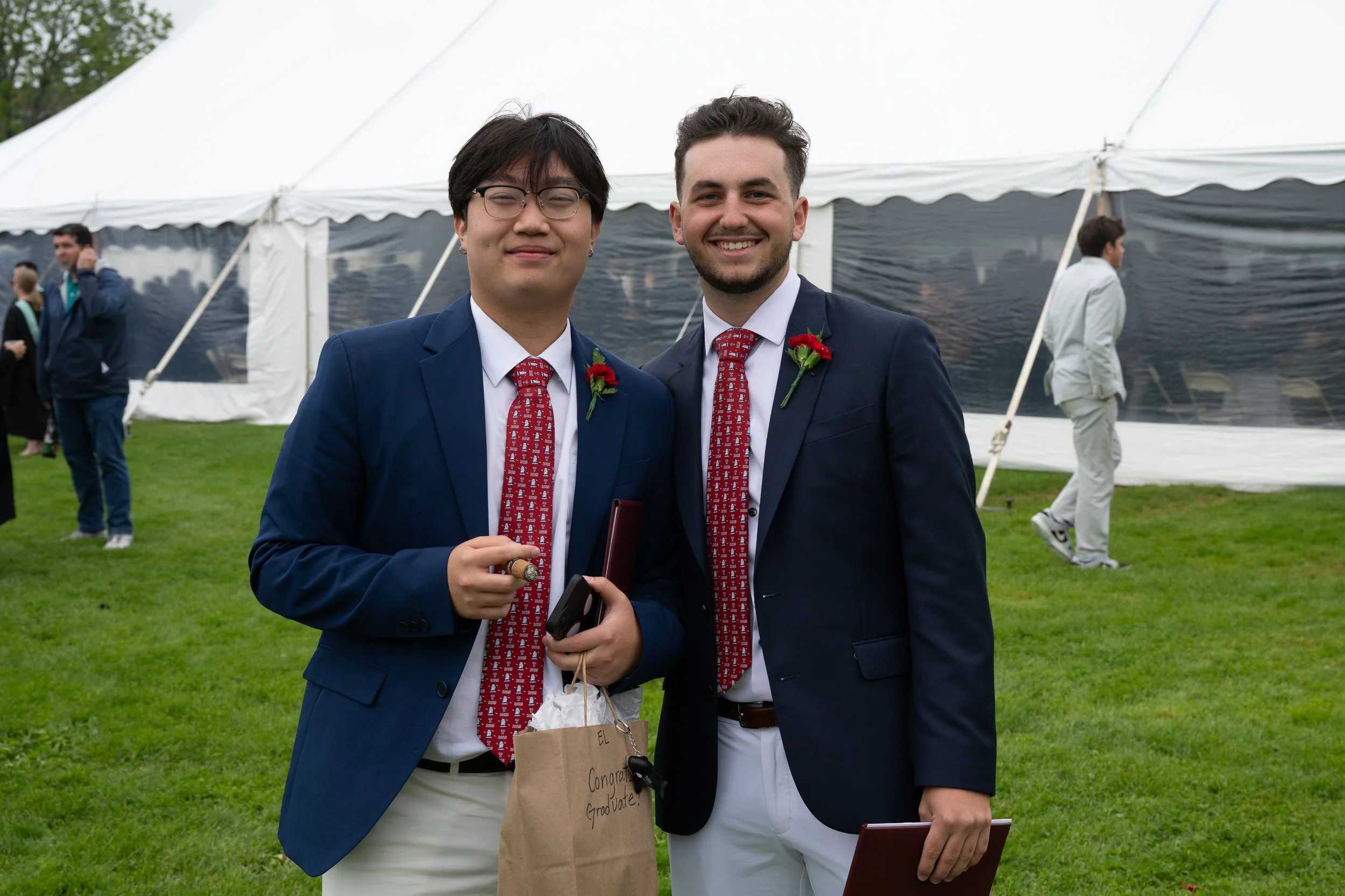 Two young men dressed in formal suits, tied with festive red ties, pose for a photo outside near a white event tent. They are smiling and holding items including a small paper bag with a congratulatory message for a graduation. Other people are visible in the background at the outdoor event.