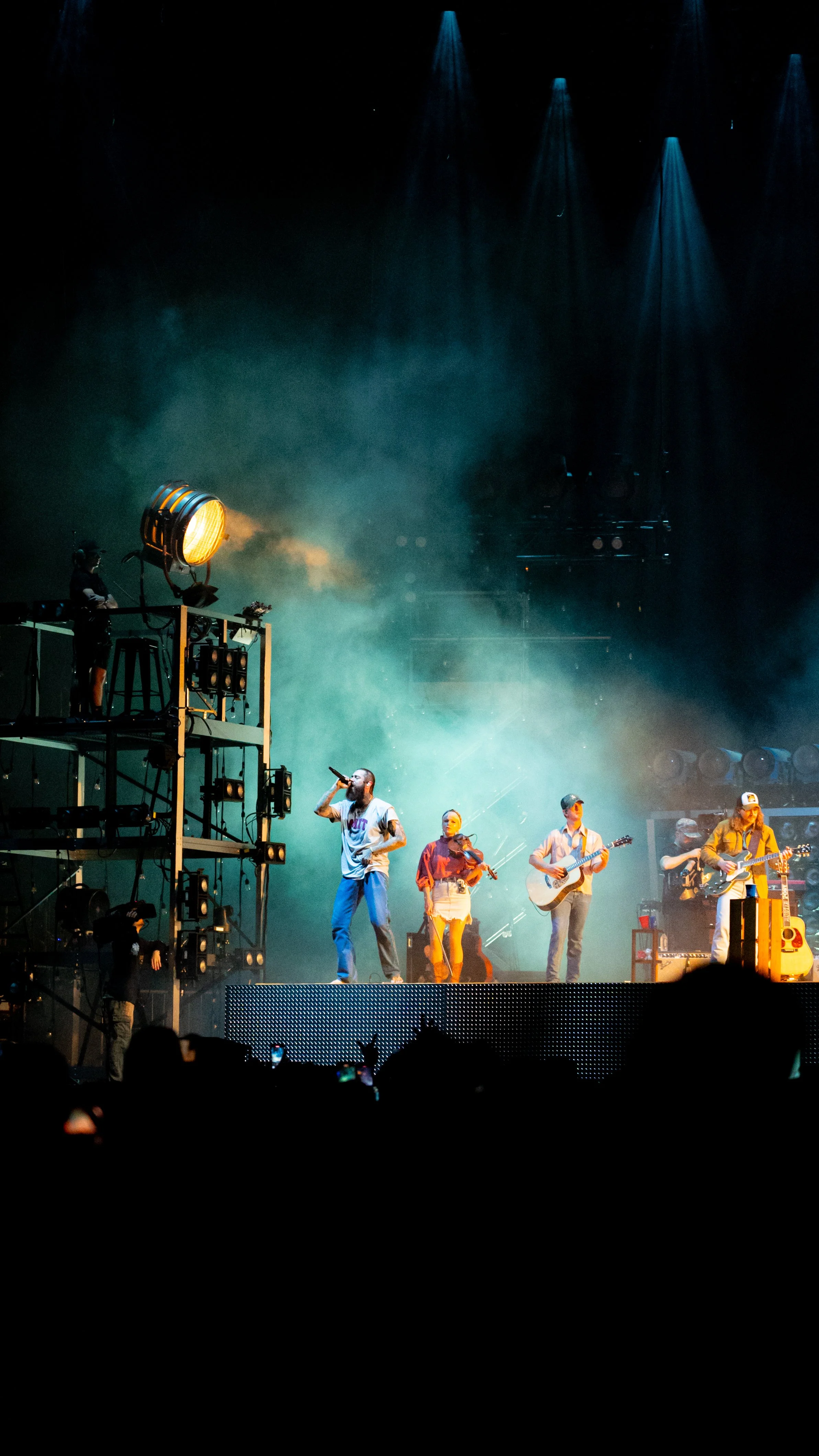 Musicians perform on stage with colorful lighting and fog, including a singer, violinist, and guitarists, during a concert.