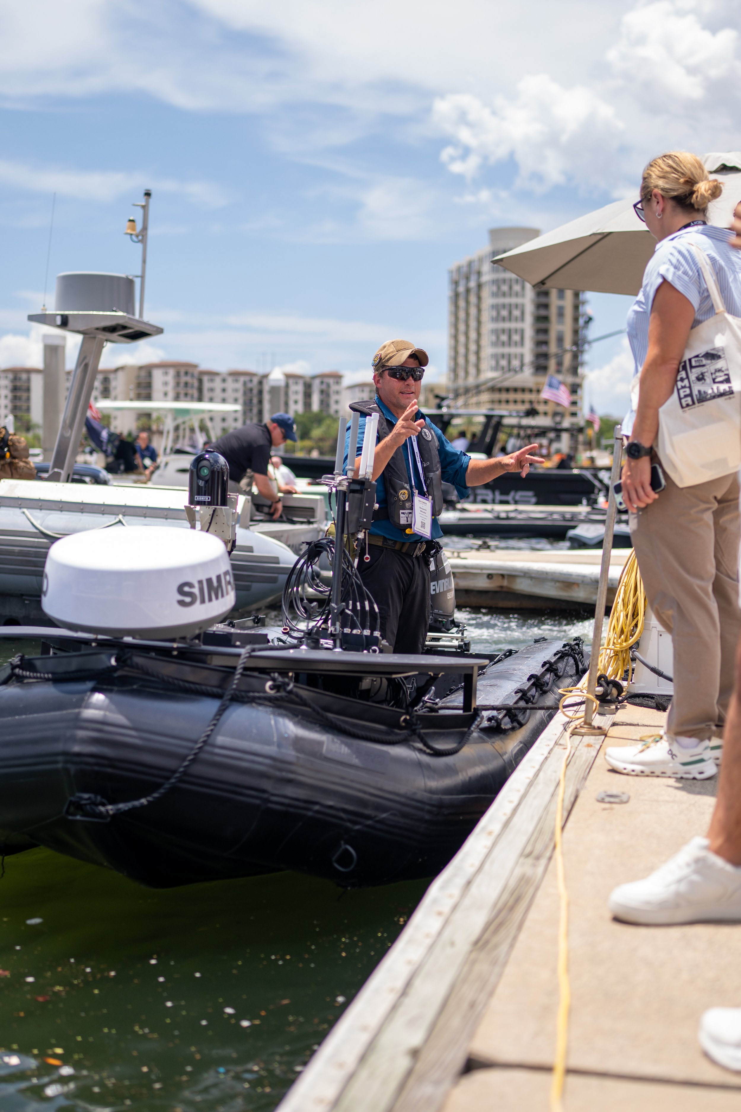 A man and woman by a dock with boats, buildings, and a cloudy sky in the background. The man is talking, gesturing with his hands, while the woman listens. The boats are equipped with electronic equipment, and there are American flags on some boats.