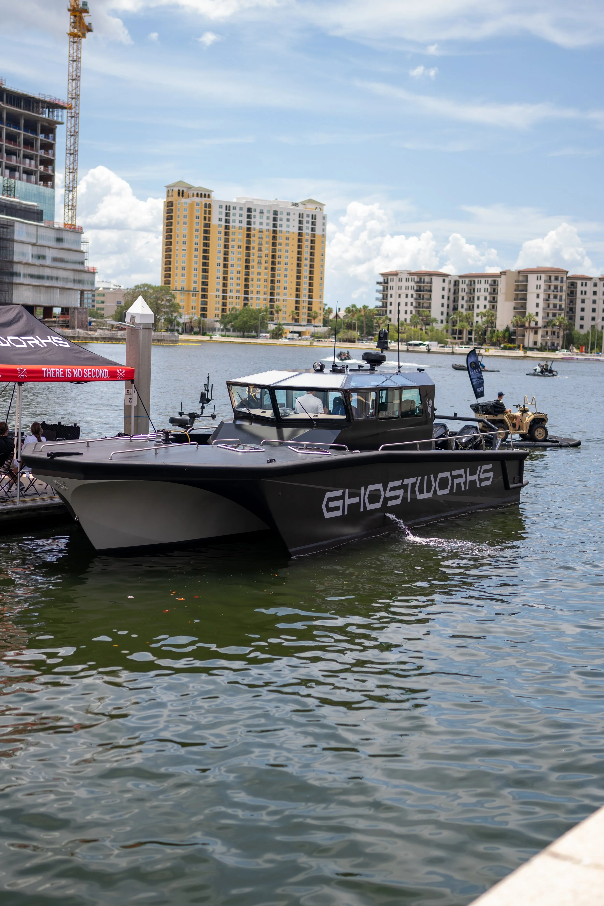 A black boat named 'GHOSTWORKS' is docked by the water, with a person inside. In the background, there are tall buildings and a construction site, with a partly cloudy sky overhead.