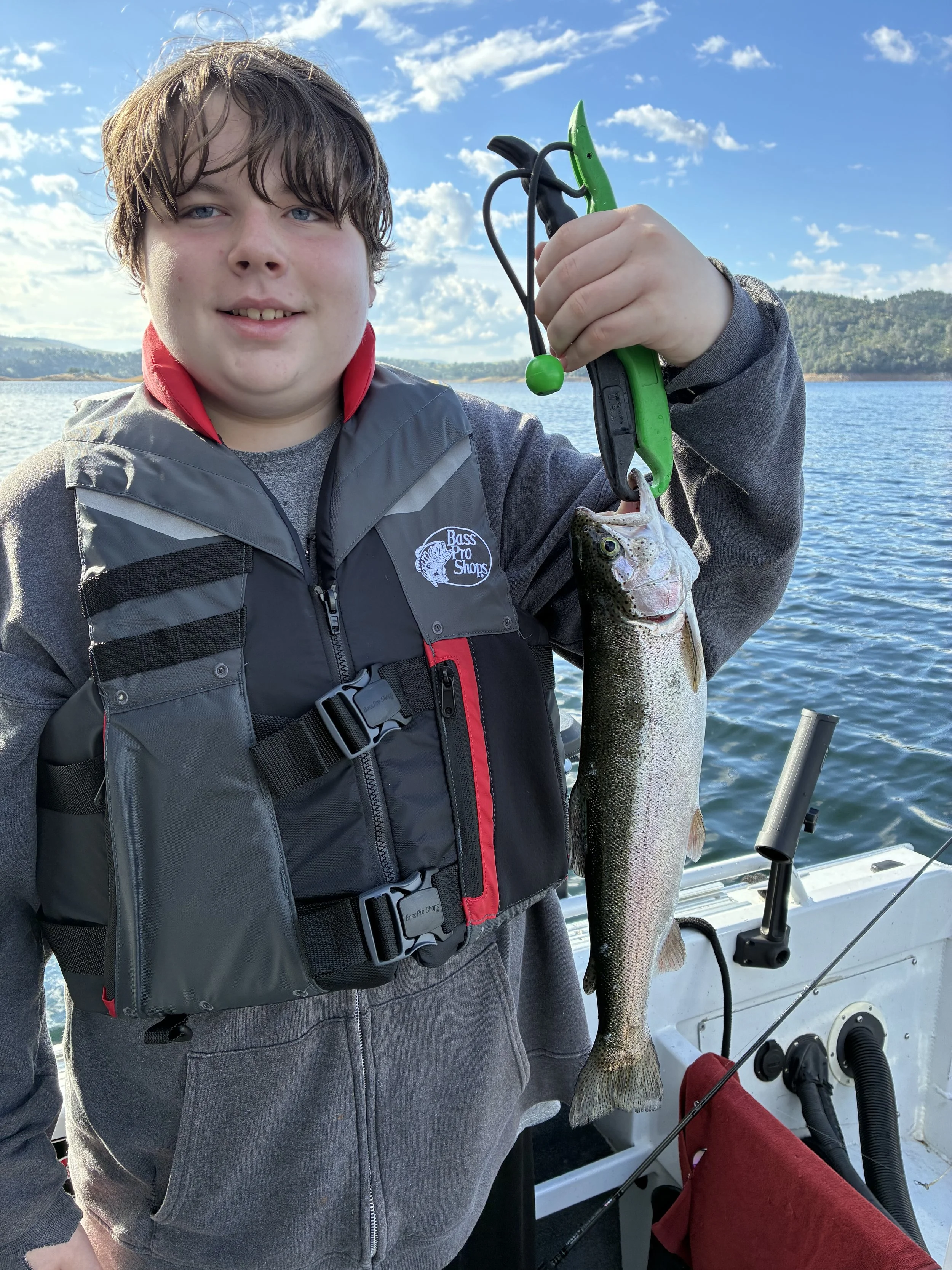 A boy with brown hair and blue eyes holding a fish on a fishing boat. The boy is wearing a gray and black vest with a red lining and a gray hoodie underneath. The fish is large, silvery with a darker back, and is hanging vertically from his hand. The