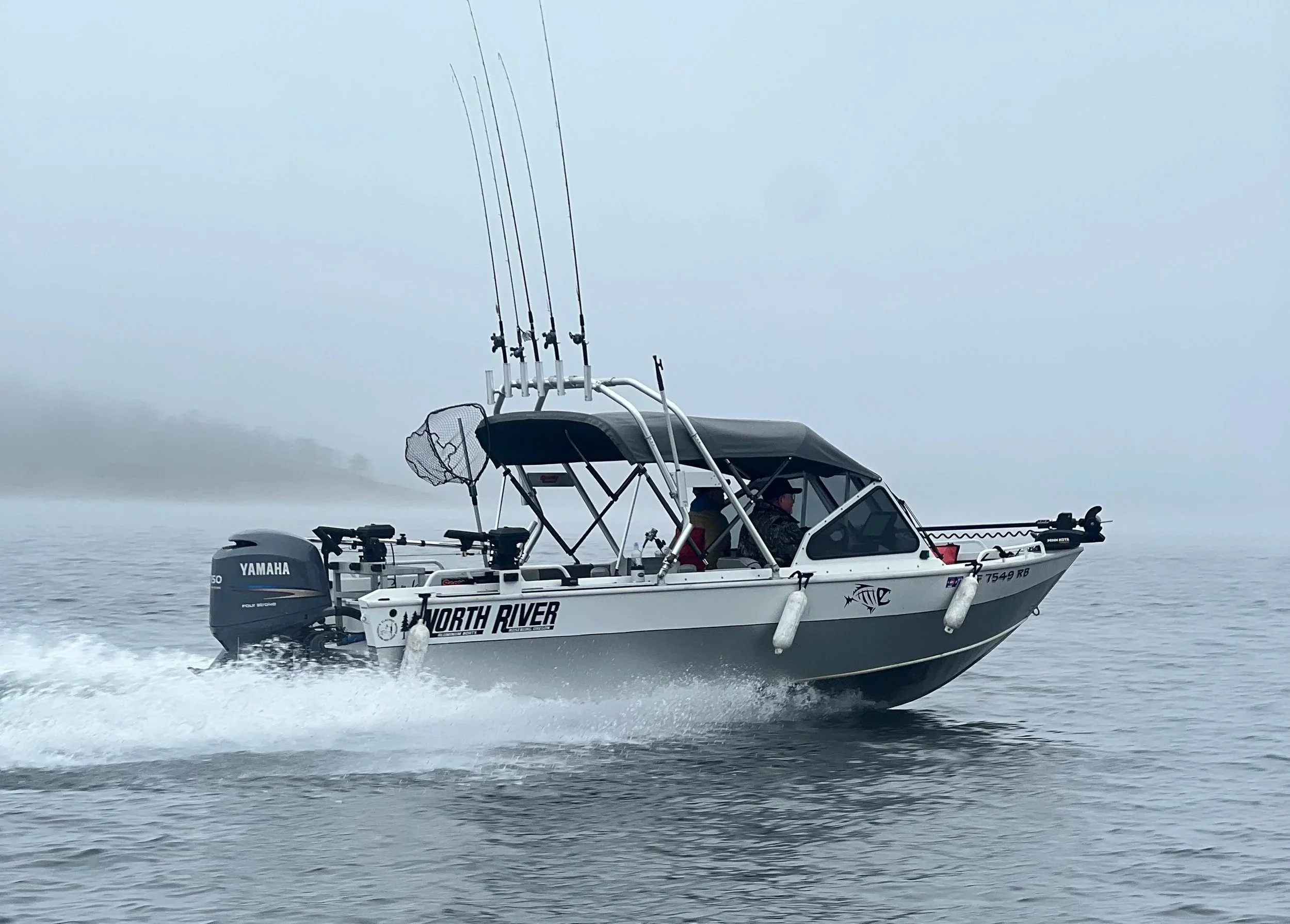 A white boat labeled North River with a Yamaha outboard motor, moving on the water with two people onboard, equipped with fishing rods and a canopy, under an overcast sky.