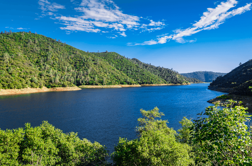 A scenic view of a river flowing between green, forested hills on a sunny day with a blue sky and scattered clouds.