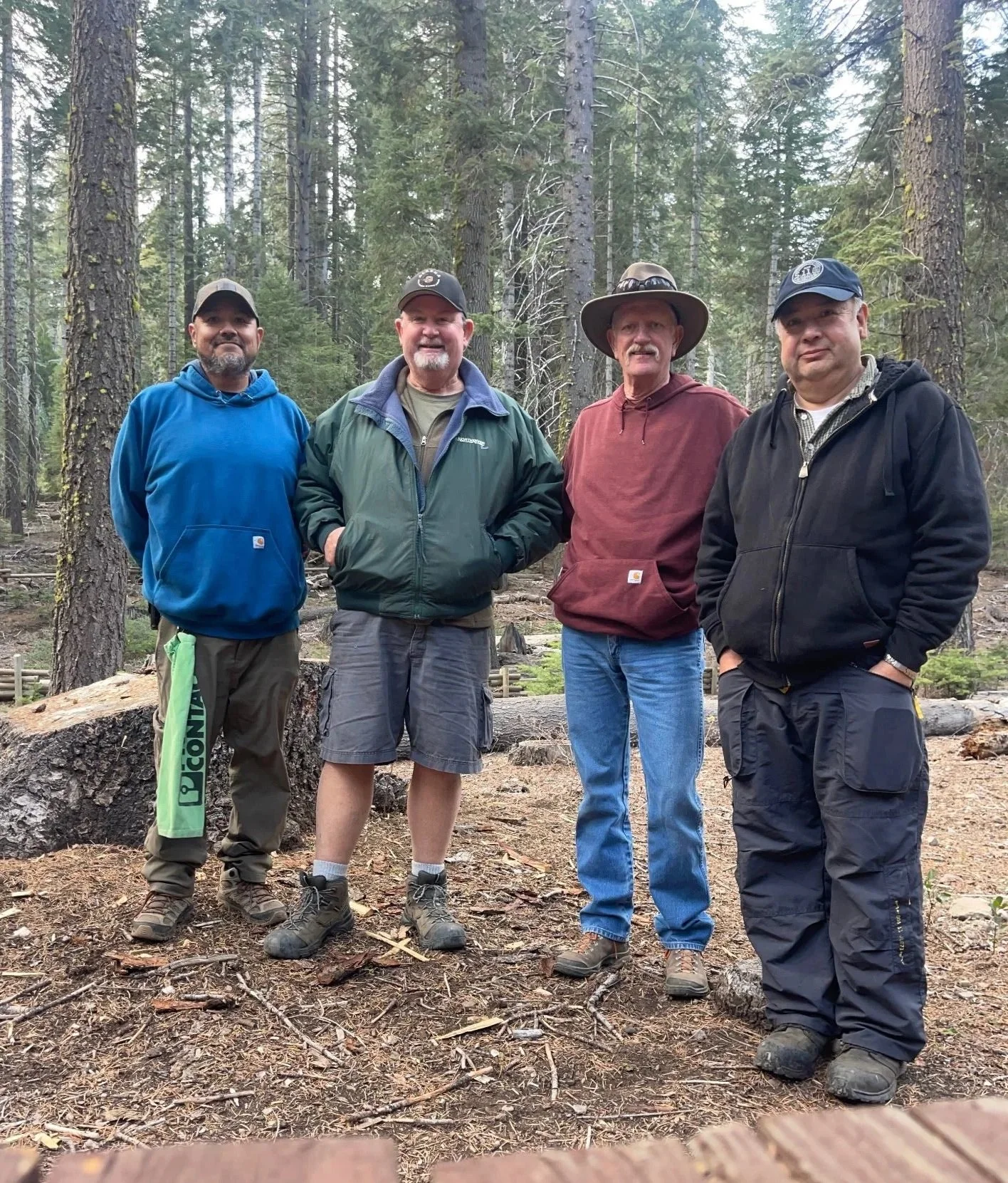 Four men standing in a forest of tall trees with moss on the trunks, wearing outdoor clothing and hiking boots.