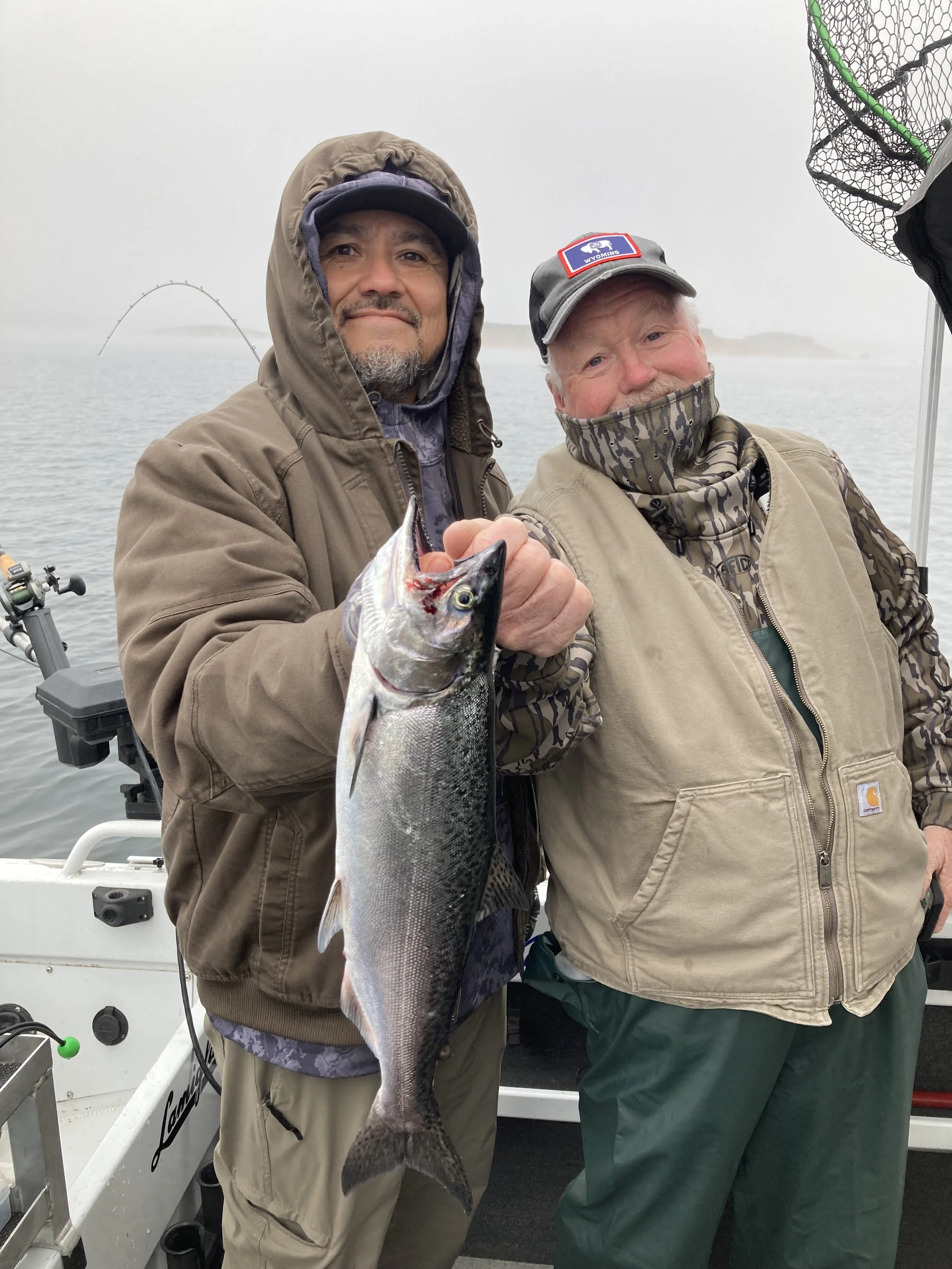 Two men on a boat holding a large fish they caught. The man on the left is wearing a brown jacket and holding the fish. The man on the right is wearing a beige vest, a gray cap with a Wyoming patch, and a camouflage neck gaiter.