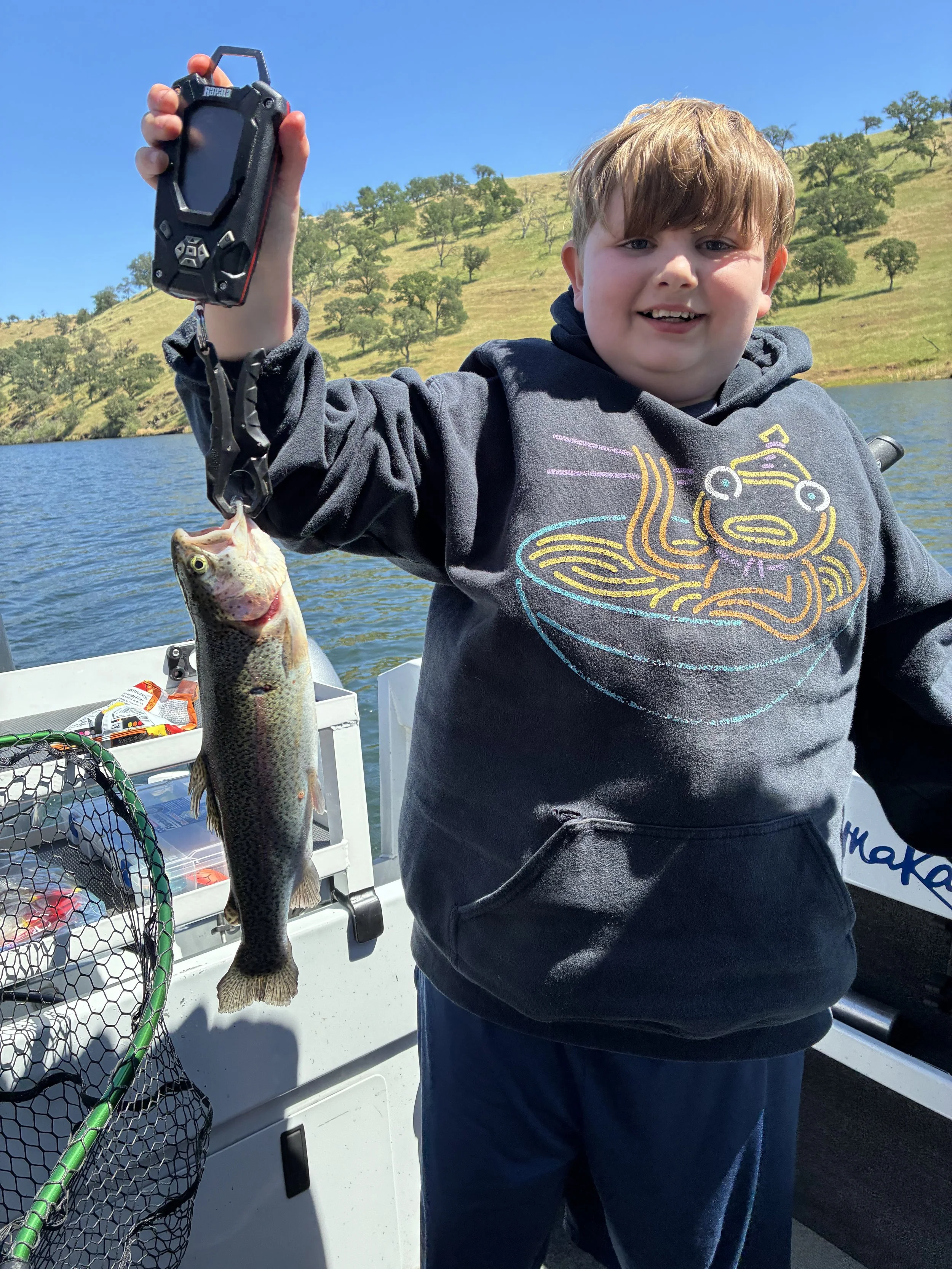 A young boy smiling and holding up a fishing rod with a small fish he caught, on a boat with a net and tackle box, with a scenic background of hills, trees, and a river under a blue sky.