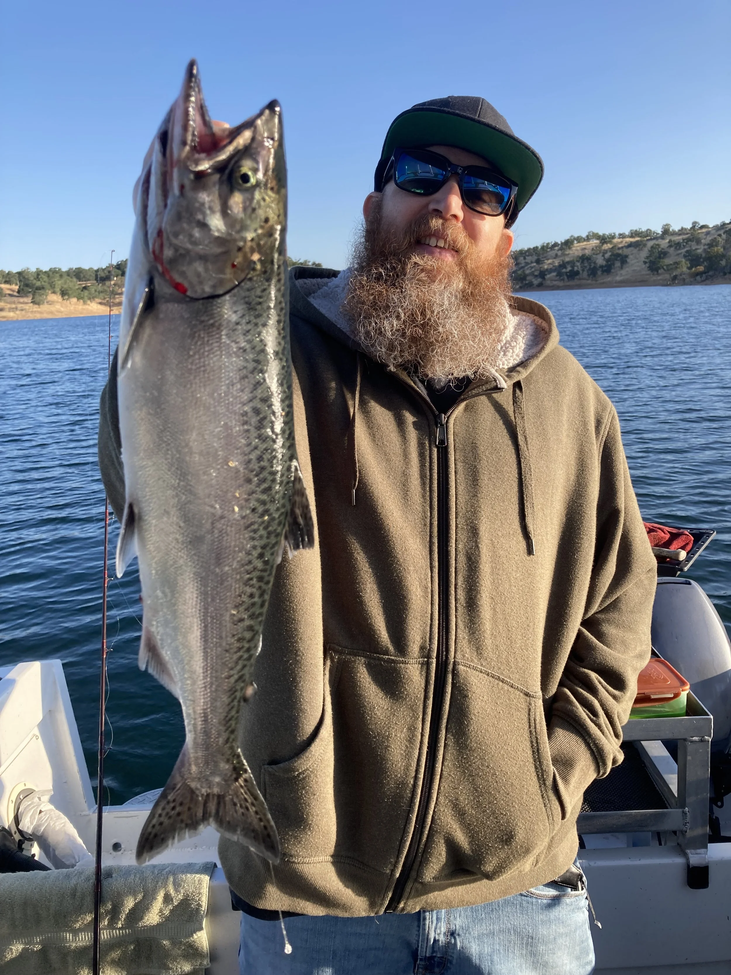 A man with a beard wearing a hoodie, sunglasses, and a cap holding a fish on a boat in a lake with hills in the background.
