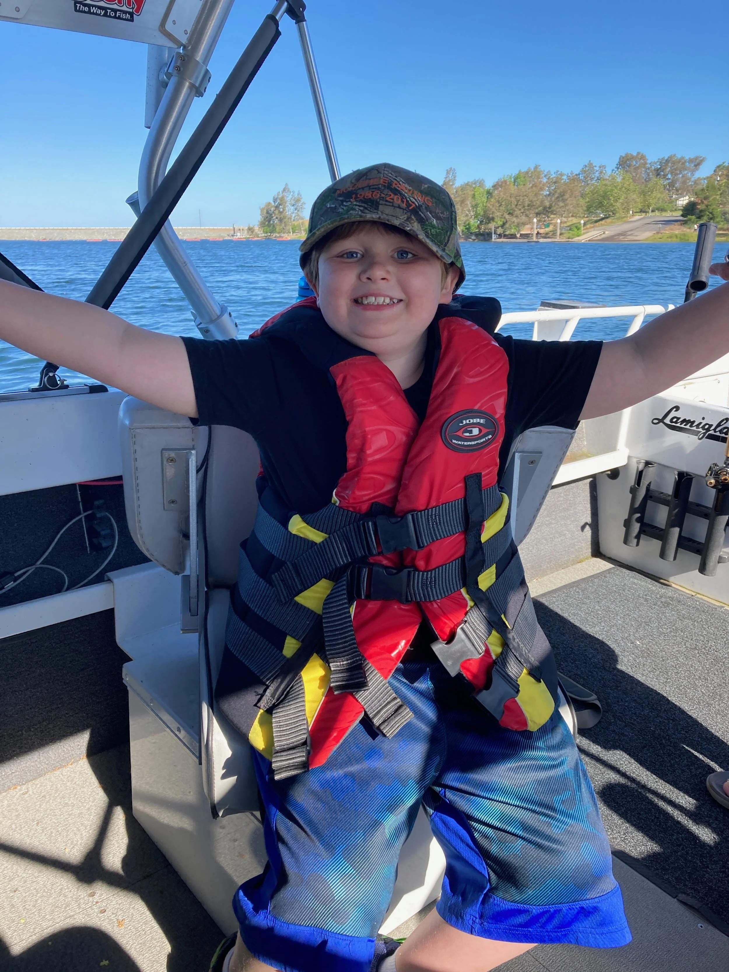 A young boy smiling on a boat with water and trees in the background, wearing a camo hat, a red life jacket, and blue shorts.
