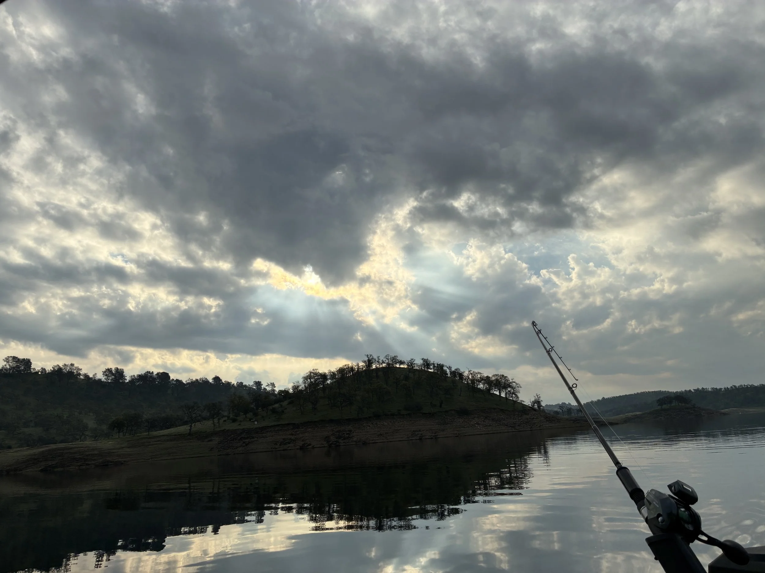 Overcast sky with clouds over a calm body of water, with tree-covered hills in the background. A fishing rod is visible in the lower right corner, leaning over the water.