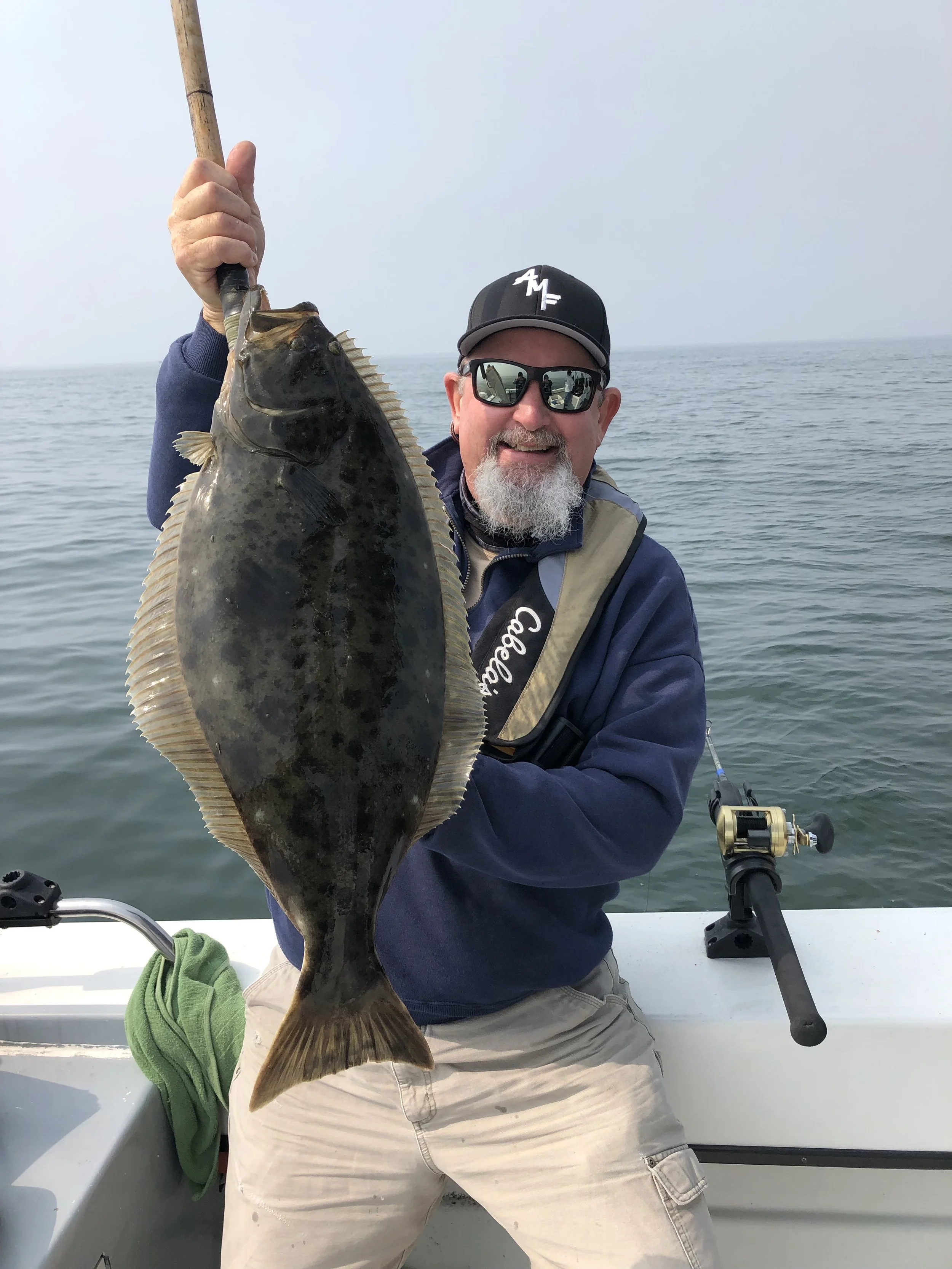 Man holding a large fish on a boat, wearing sunglasses, a baseball cap, and a fishing vest, with a fishing rod nearby, on open water with a cloudy sky.