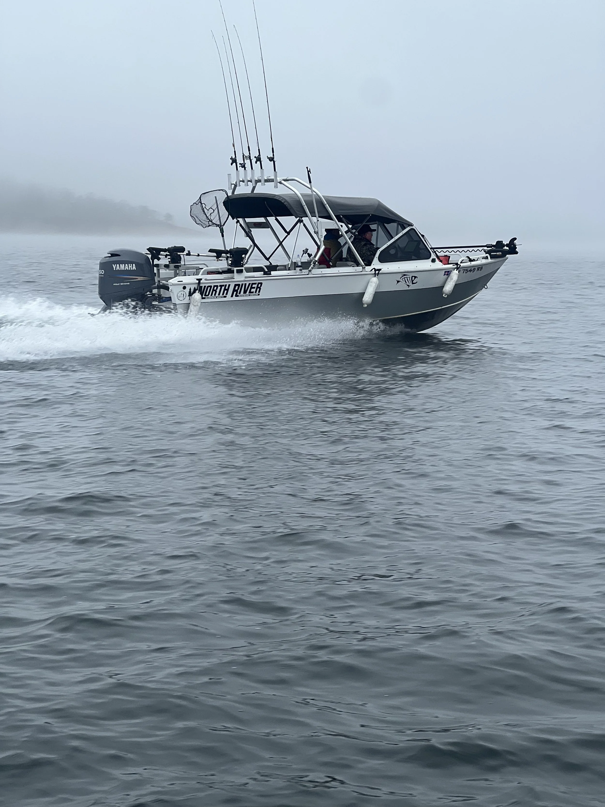 A motorboat with fishing rods riding on calm water with foggy background and some land in the distance.