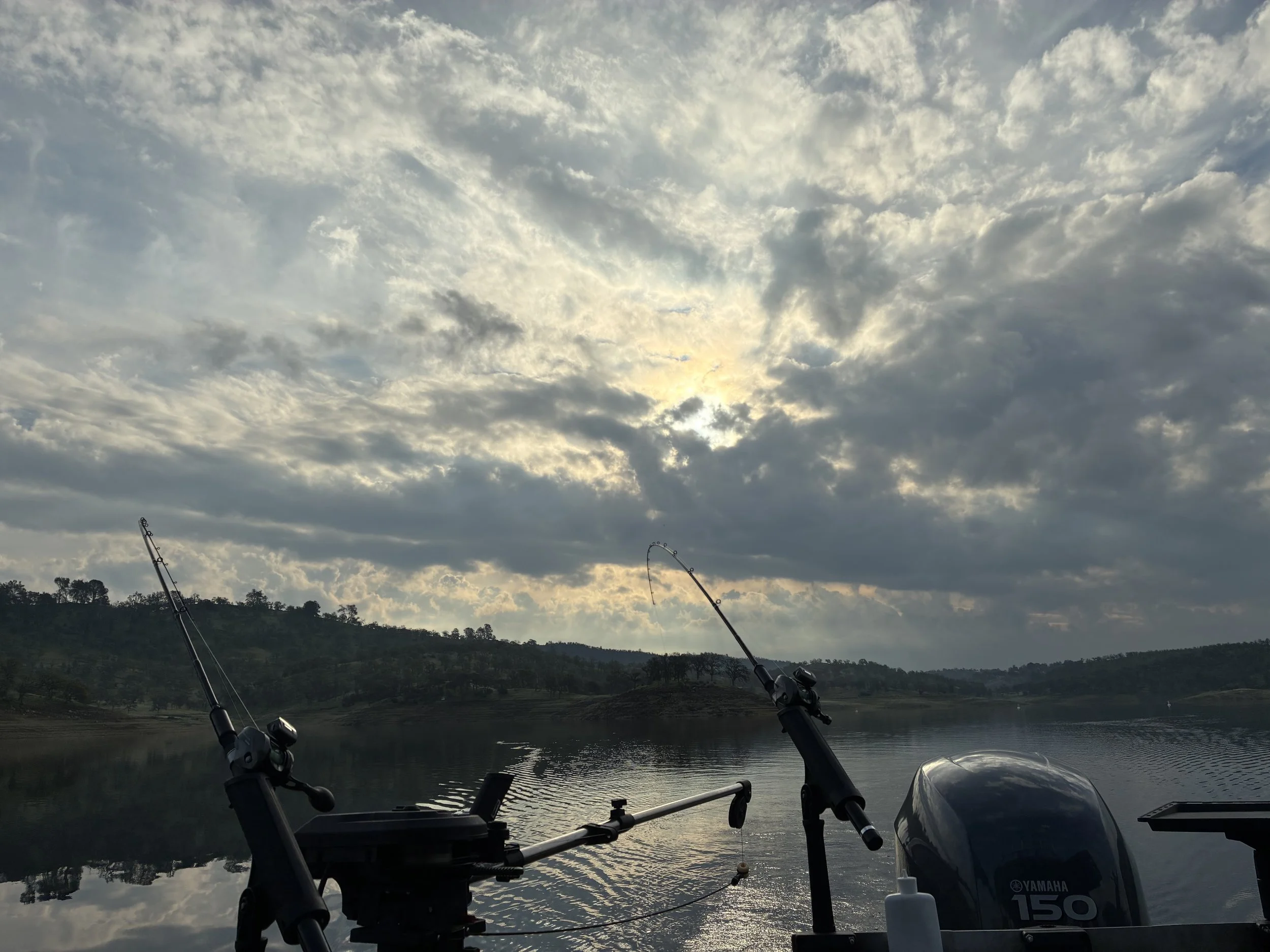 View from a boat on a lake with fishing rods in the foreground, a hilly landscape in the background, and a cloudy sky with sunlight peeking through.