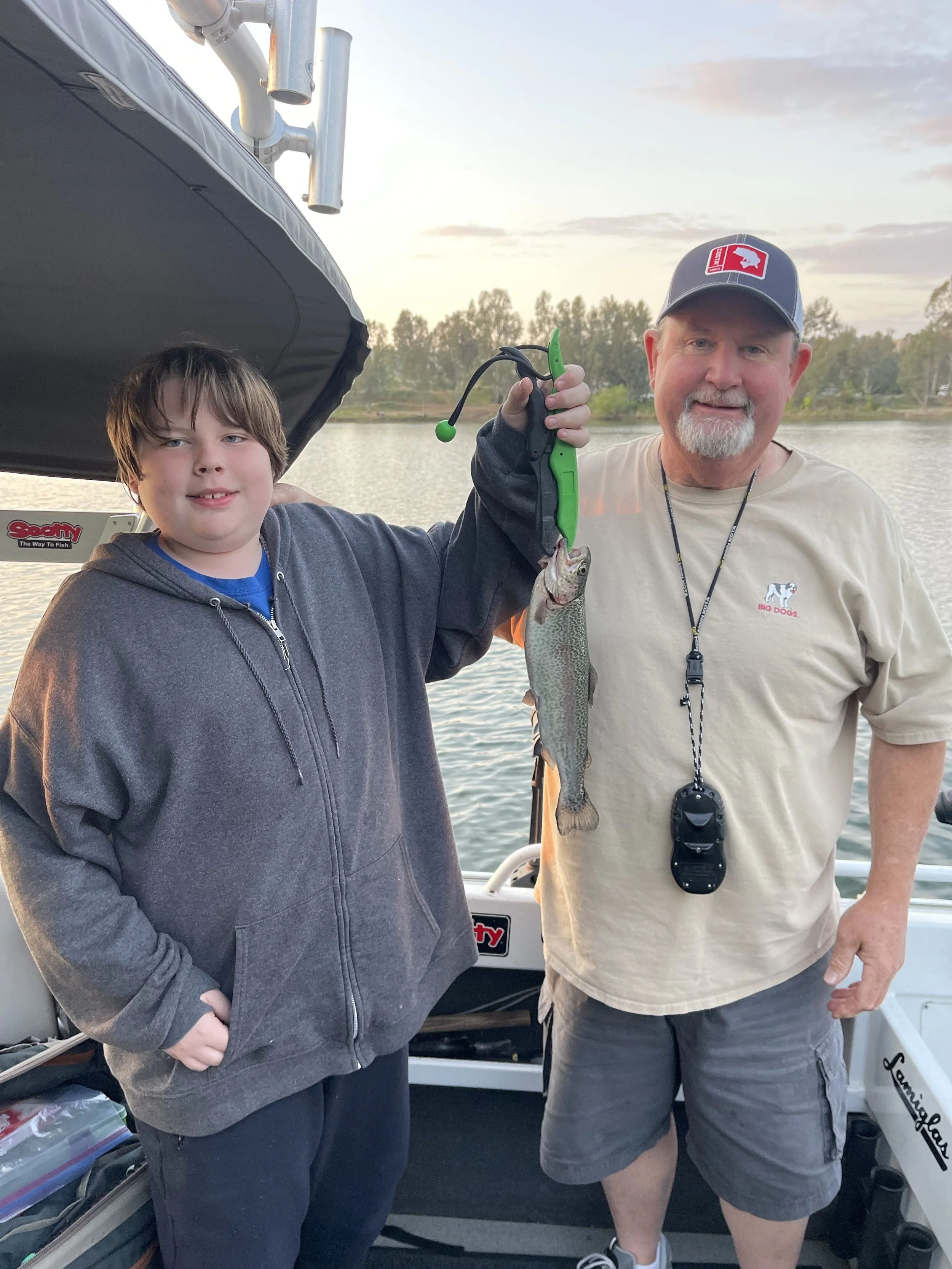 A boy and an older man on a boat holding a fish they caught together, smiling. The boy wears a gray hoodie, and the man wears a beige shirt and a gray cap. The background shows water and trees, with a sunset sky.