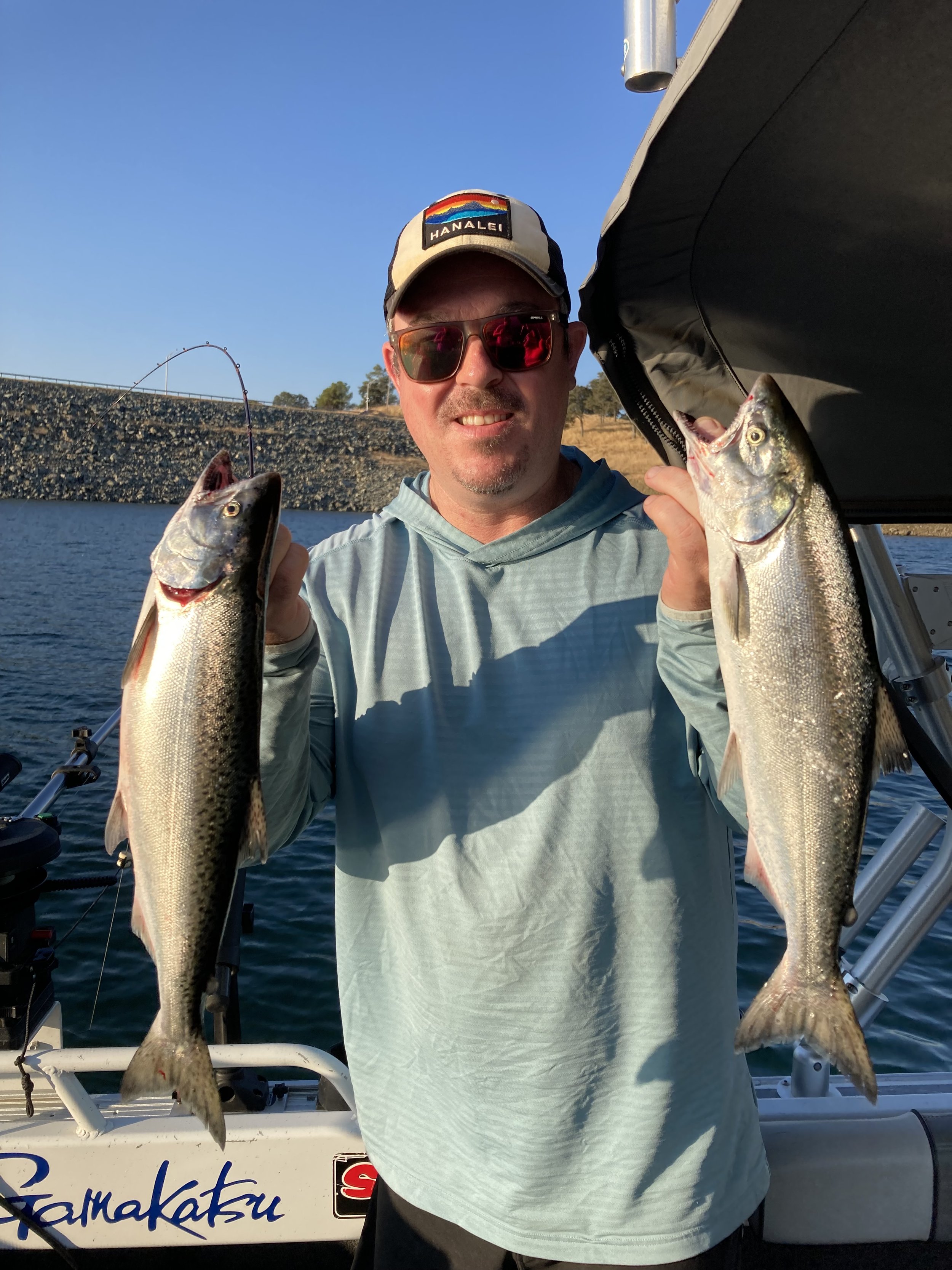Man holding two fish on a boat with water and shoreline in the background during daytime.