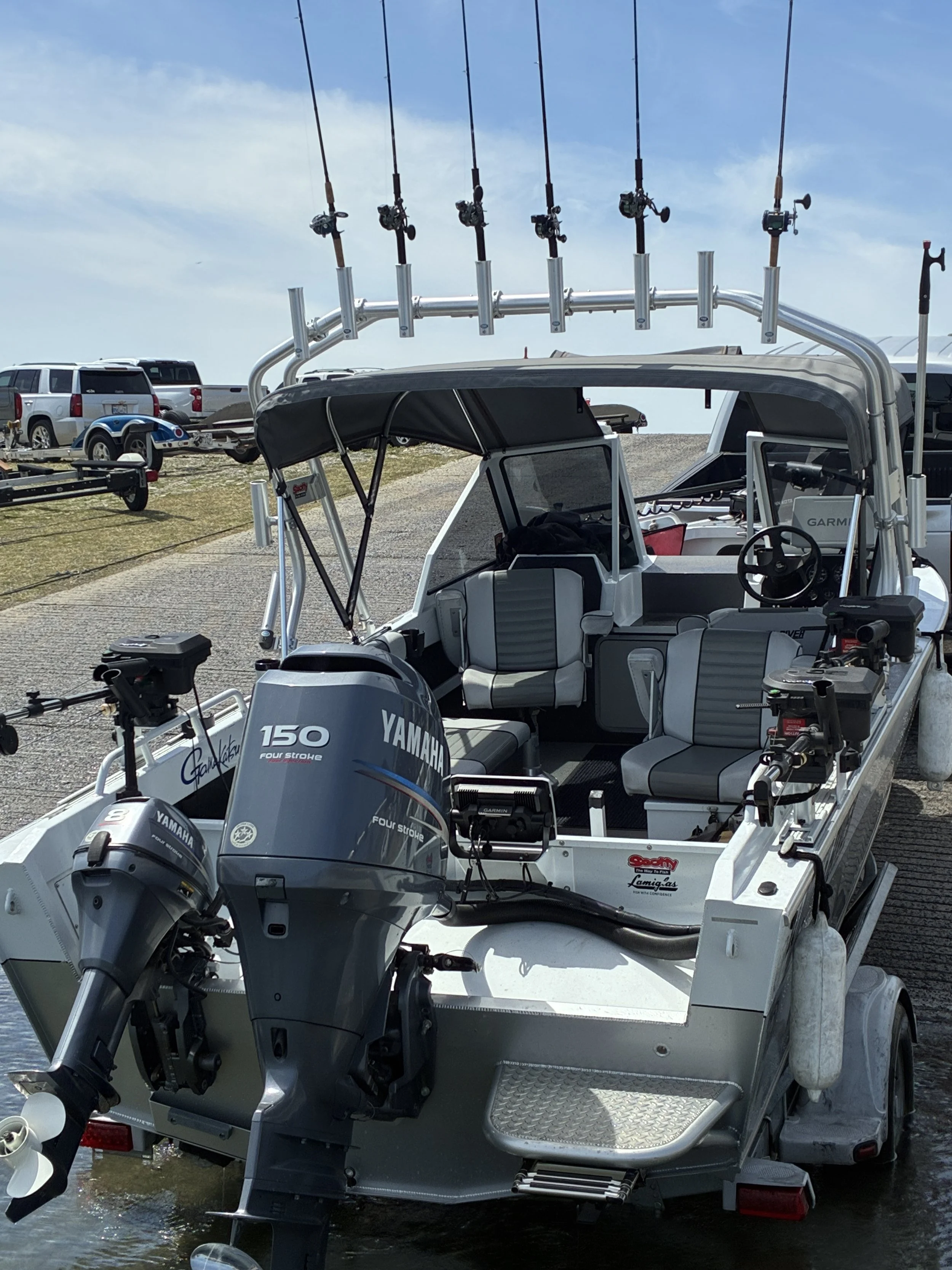 A boat with fishing rods mounted on a tower, equipped with Yamaha four-stroke outboard motor, parked on a trailer near a parking lot with several cars and a trailer in the background.