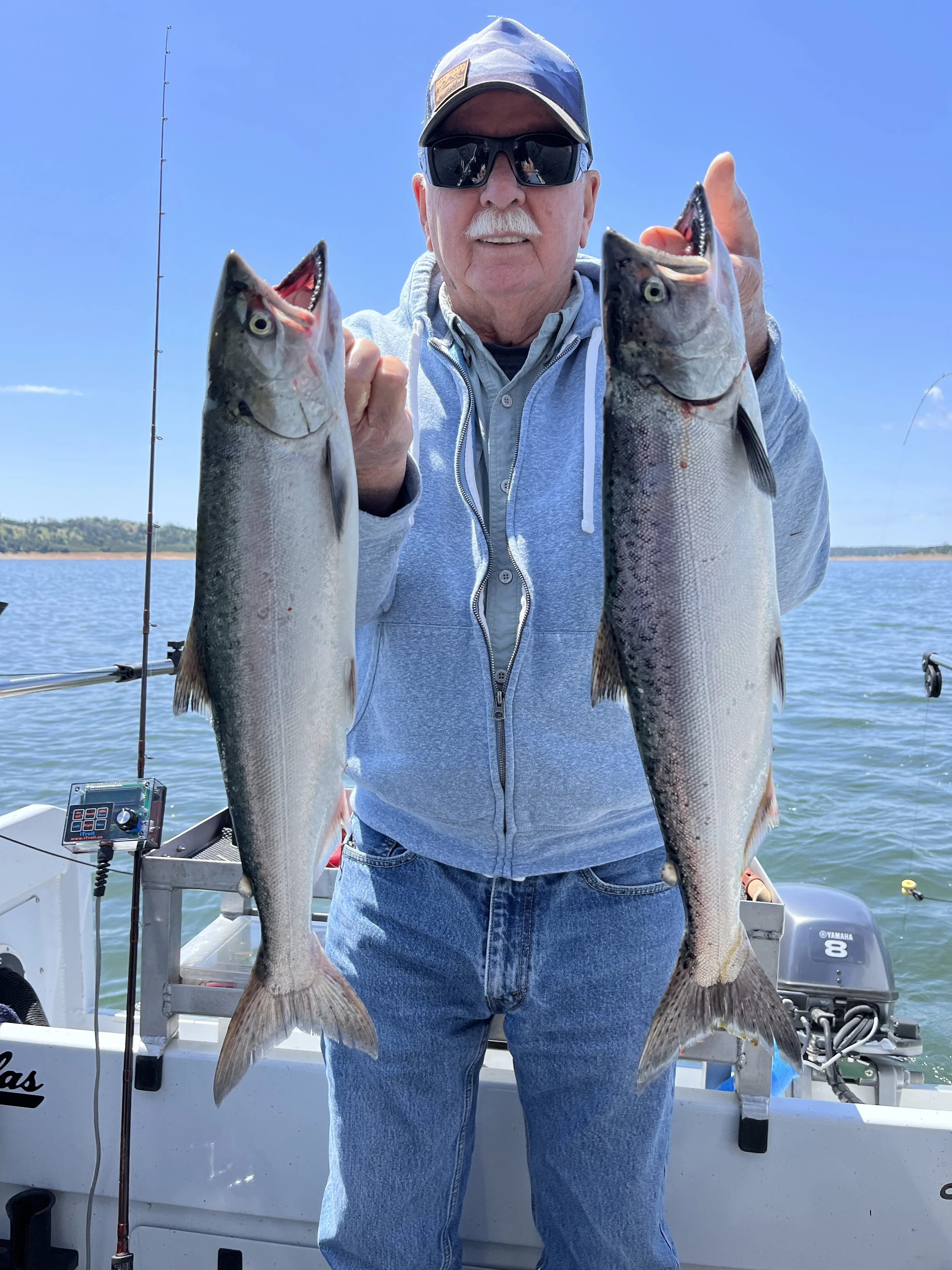 An elderly man wearing sunglasses, a cap, and a gray hoodie holding two large fish on a boat during the daytime with a clear blue sky and water in the background.