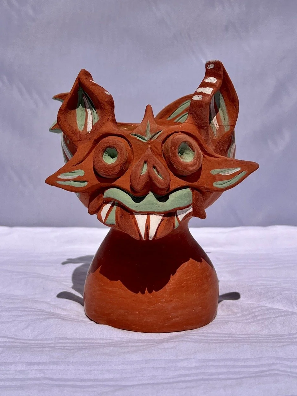 Ceramic figurine of a whimsical fox with exaggerated features, painted in orange, green, and white, on a white surface with a gray background.