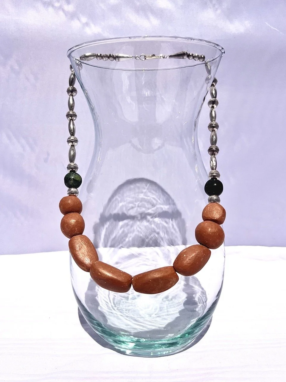 A glass vase with a beaded necklace draped over its rim, featuring orange, black, green, and silver beads, with the necklace hanging inside the vase and casting reflections on the glass surface.