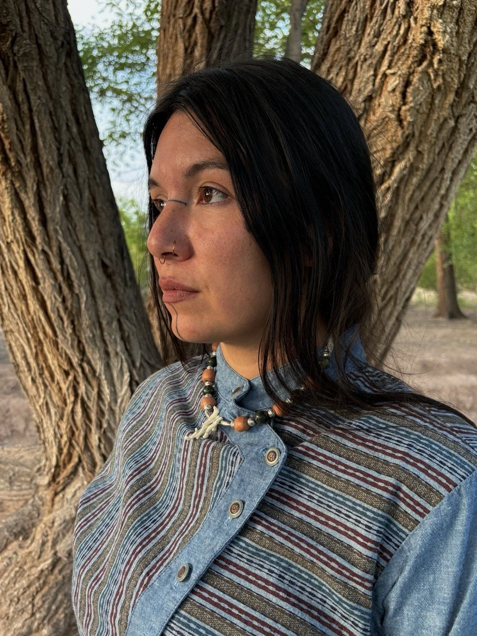 A woman with black hair, a nose ring, and wearing a striped shirt and beaded necklace standing outdoors near a tree.