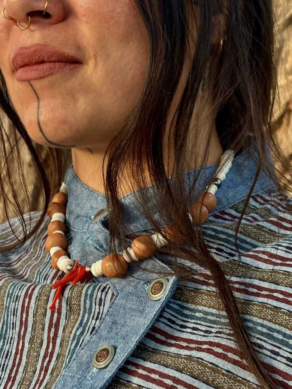 Close-up of a person with dark hair, visible lower face, and neck, wearing a beaded necklace, earring, and a colorful striped shirt with buttons.