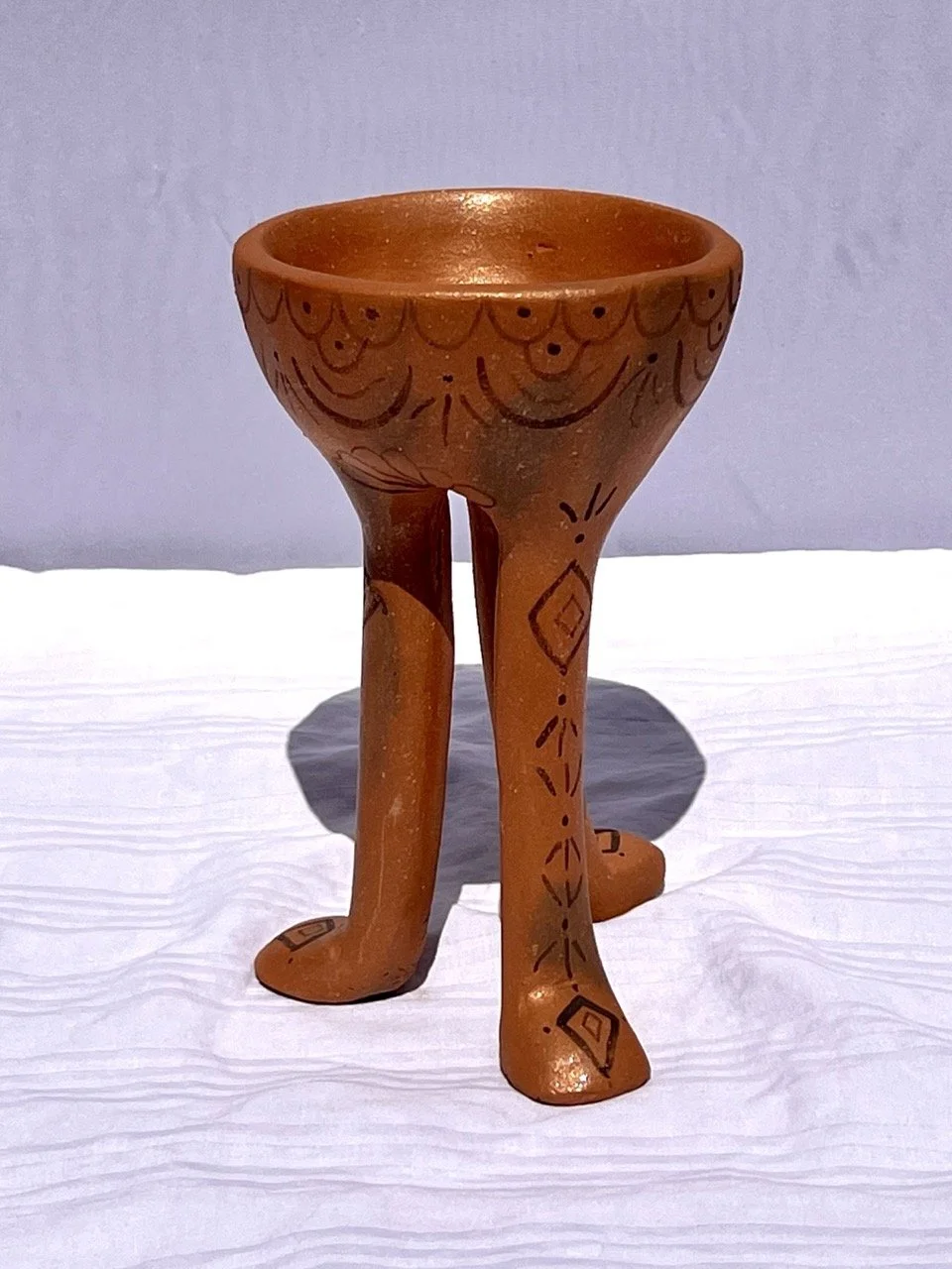 A small terracotta sculpture with a bowl on top supported by three legs that resemble animal legs, decorated with black geometric and floral designs.