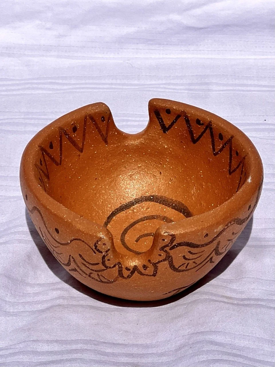 A small, handcrafted, orange clay bowl with decorative black painted designs, including zigzag patterns and a spiral, placed on a white textured surface.