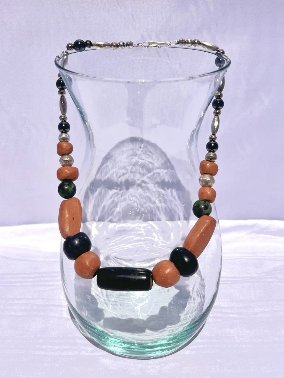 A beaded necklace displayed on the rim of a clear glass vase with a rounded body, against a white background.