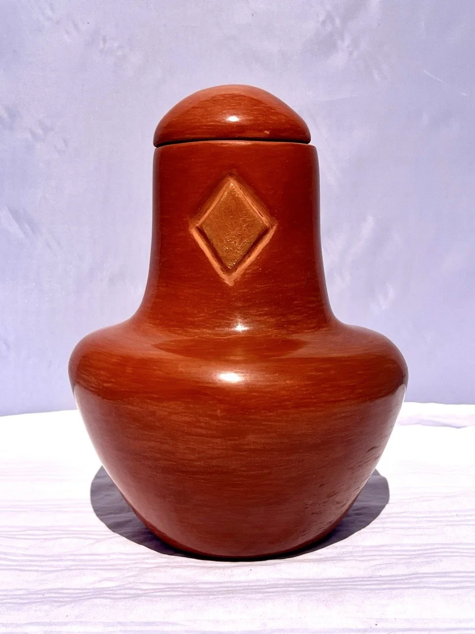A polished wooden jar with a diamond-shaped carving on its front, placed on a white surface against a light-colored wall.