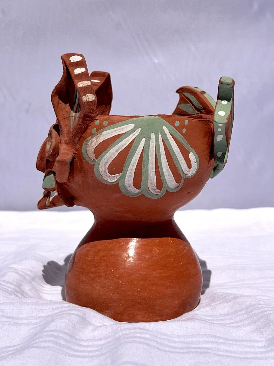 A handcrafted ceramic vessel with decorative painted patterns in white and green on an orange background, set against a white surface and background.