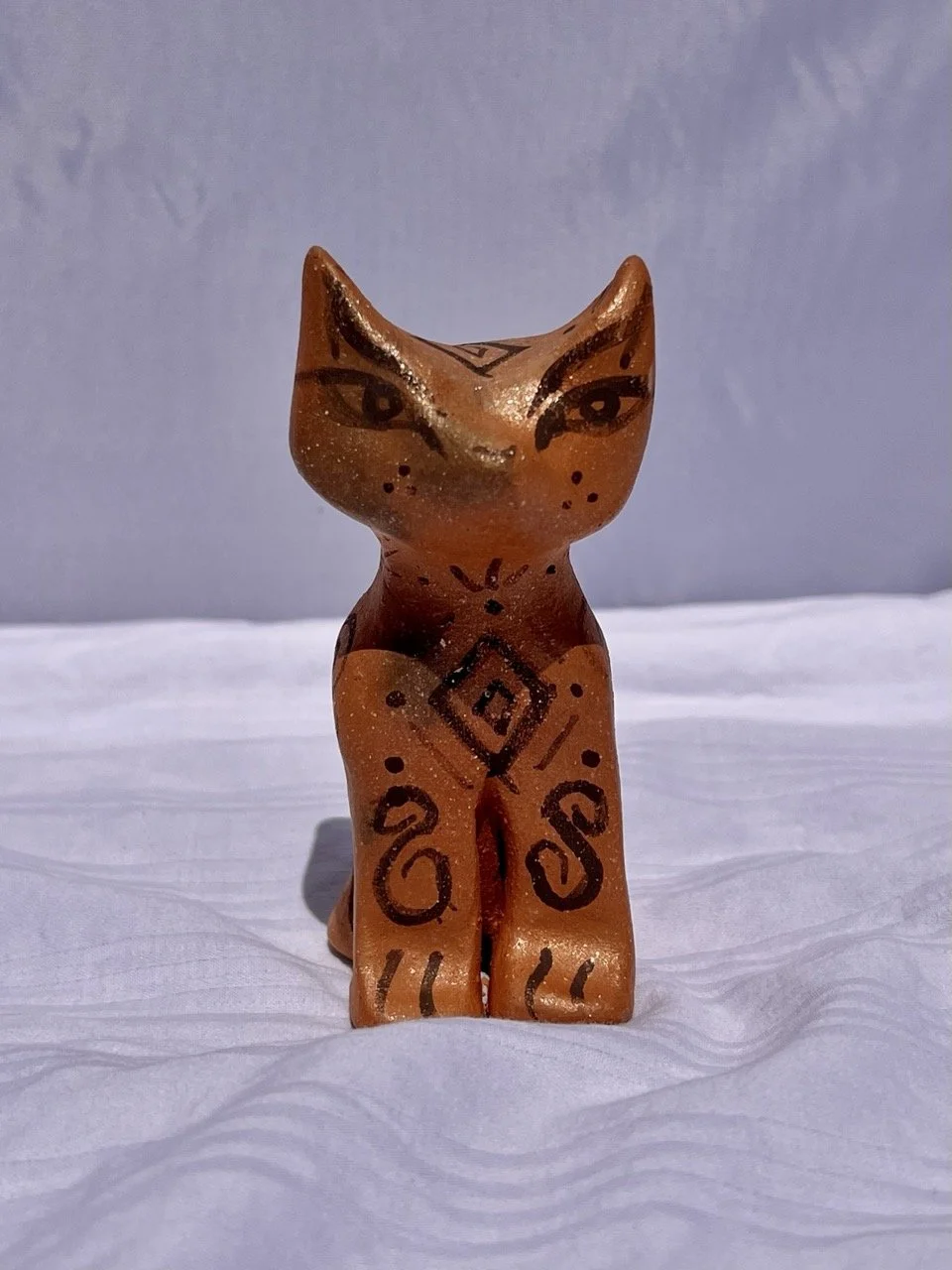 A small figurine of a stylized cat with painted facial features and decorative patterns on its body, placed on a white textured surface against a plain background.