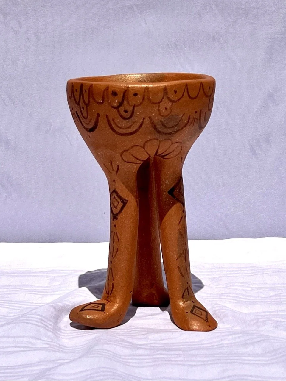A ceramic sculpture of a bowl with two human legs as the base, decorated with hand-painted designs, set on a white surface with a plain background.