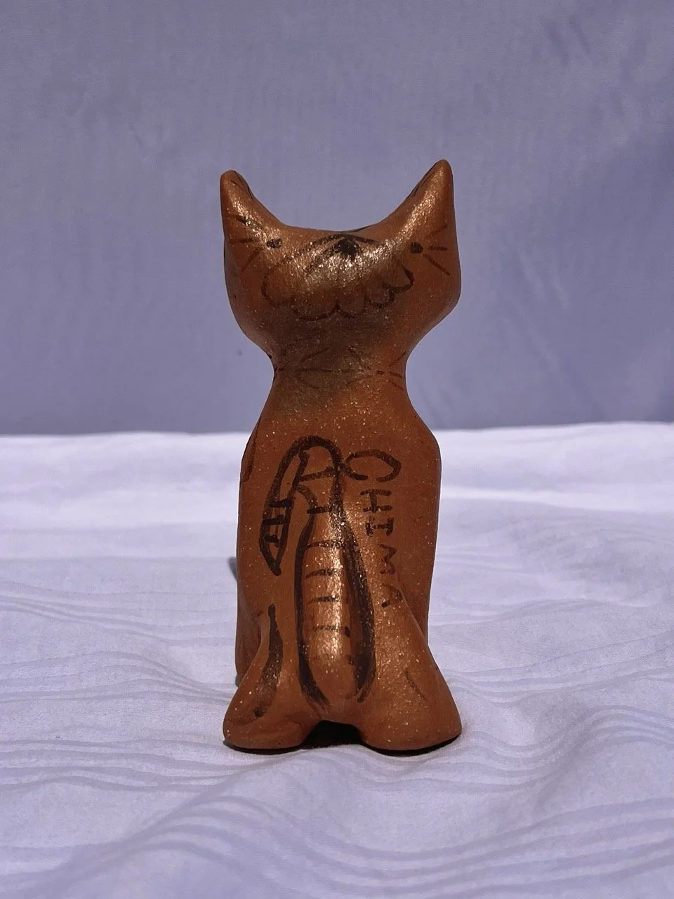 Small terracotta figurine of a cat with painted facial features and markings, placed on a white textured surface.