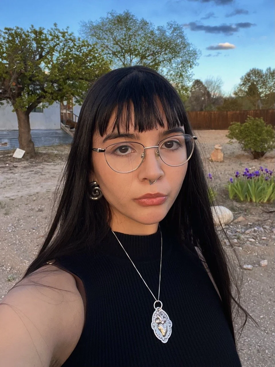 A woman with long dark hair, glasses, and a septum piercing takes a selfie outdoors during sunset. She wears a black sleeveless top and a silver pendant necklace, standing in a garden with trees and purple flowers.