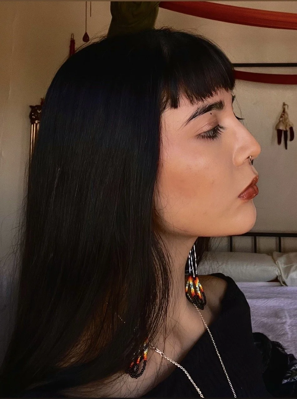 Profile of a woman with long, dark hair and bangs, wearing colorful beaded earrings and a necklace, in a dimly lit room with a bed and decorative hanging items in the background.