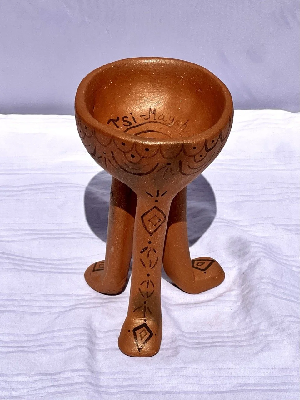 Clay bowl with decorated legs, displaying the name 'Rsi-May' inside, placed on a white surface.