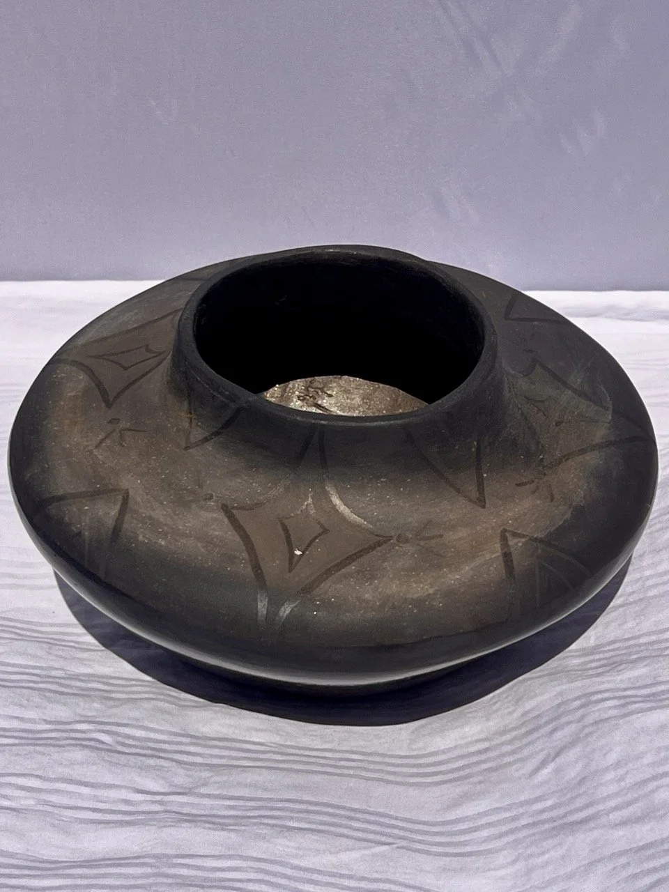 A black, rounded ceramic vessel with geometric patterns etched on its surface, placed on a light-colored wooden surface.
