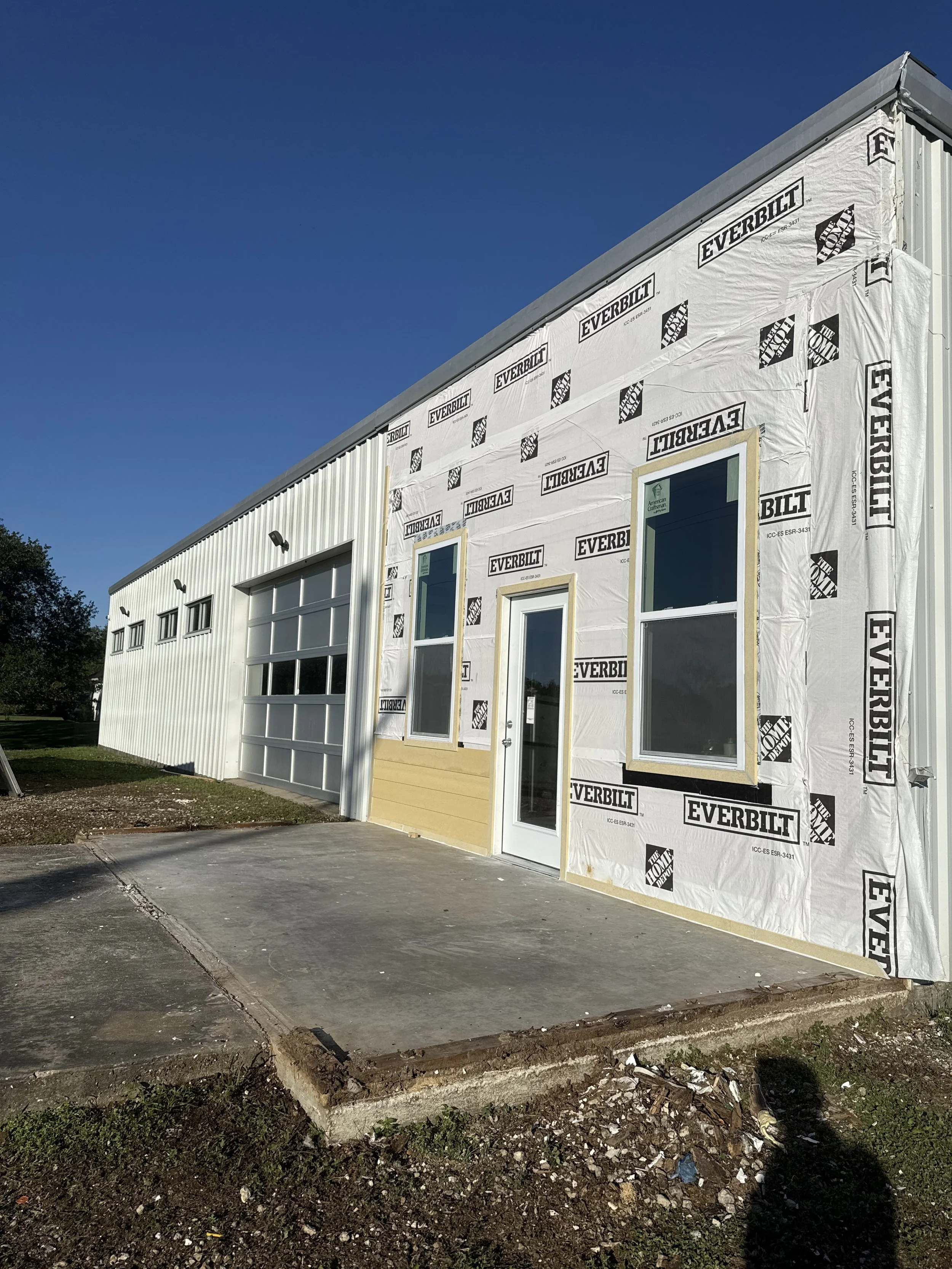 Building under construction with white everbilt house wrap, three windows, and a garage door, with a concrete sidewalk in front, and a blue sky overhead.