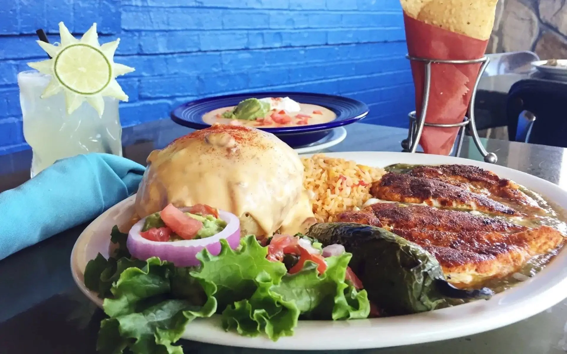 Plate with Mexican food including chicken topped with cheese sauce, rice, grilled chicken, roasted peppers, guacamole, and fresh salad; background shows blue wall and drink with a lemon slice and sun-shaped lemon decoration, with some items on the table.