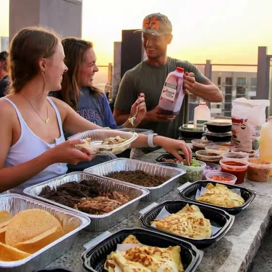 People enjoying a patio barbecue with various dishes, including tacos, pizza, and grilled meats, on a rooftop at sunset.