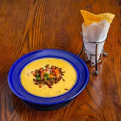 A bowl of yellow soup garnished with chopped vegetables and herbs, placed on a wooden table, with a glass holder containing a paper napkin and yellow tortilla chip on the side.